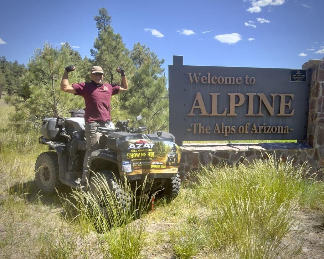 Rusty Childress raises his arms beside his ATV at the "Welcome to Alpine" trailhead sign, marking the start of his 1,000-mile Arizona Alpine Trail ambassador ride.